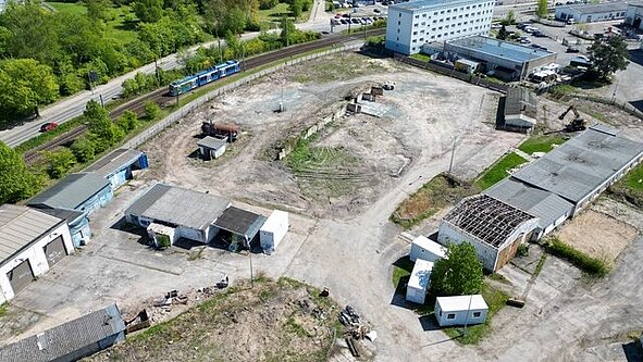 Luftaufnahme des großen, unbebauten Baugrundstücks mit einigen alten Gebäuden und Baumaschinen, umgeben von Grünflächen und dem nahegelegenen Wohngebiet Dierkow