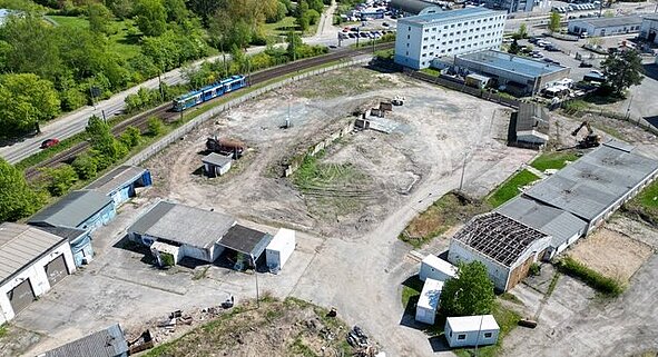 Luftaufnahme des großen, unbebauten Baugrundstücks mit einigen alten Gebäuden und Baumaschinen, umgeben von Grünflächen und dem nahegelegenen Wohngebiet Dierkow