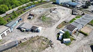 Luftaufnahme des großen, unbebauten Baugrundstücks mit einigen alten Gebäuden und Baumaschinen, umgeben von Grünflächen und dem nahegelegenen Wohngebiet Dierkow