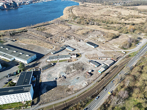 Luftaufnahme des Geländes des künftigen WarnowQuartiers mit Blick auf die ungenutzten Gebäude des ehemaligen Bauhofes, umgeben von Wasser und Straßen. Im Hintergrund sind Gebäude und eine Stadt sichtbar.