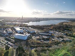 Panoramablick auf das WarnowQuartier mit Wasser im Hintergrund, umgeben von Hügeln und Gebäuden. Die Sonne scheint hell und reflektiert auf der Wasseroberfläche.