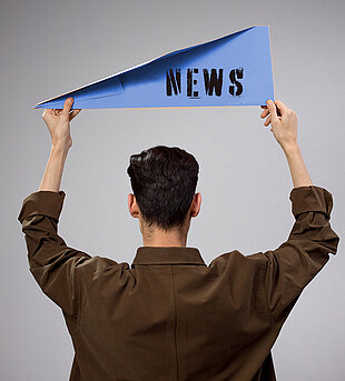 Person von hinten, die ein blaues Schild mit der Aufschrift 'NEWS' hält, trägt ein braunes Oberteil.