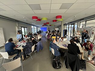 Gruppendiskussion in einem modernen Raum mit Tischen und Stühlen, Teilnehmer sitzen in einem Kreis, bunte Lampen hängen von der Decke.