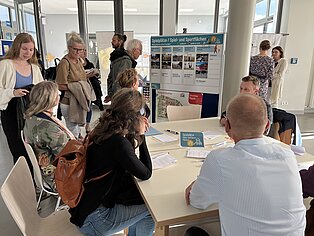 Gruppe von Menschen in einem modernen Raum, die an einem Tisch sitzen und diskutieren. Im Hintergrund sind Plakate und Informationsmaterialien sichtbar.