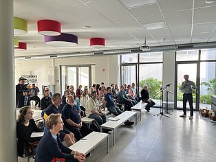Eine Präsentation in einem modernen Raum mit großen Fenstern. Eine Gruppe von Menschen sitzt auf weißen Bänken und hört einem Redner zu, der vorne steht.