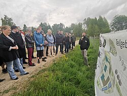 Gruppe von Menschen, die im Freien vor einem Plakat stehen. Ein MAnn hält ein Mirkofon und erklärt etwas auf dem Plakat.
