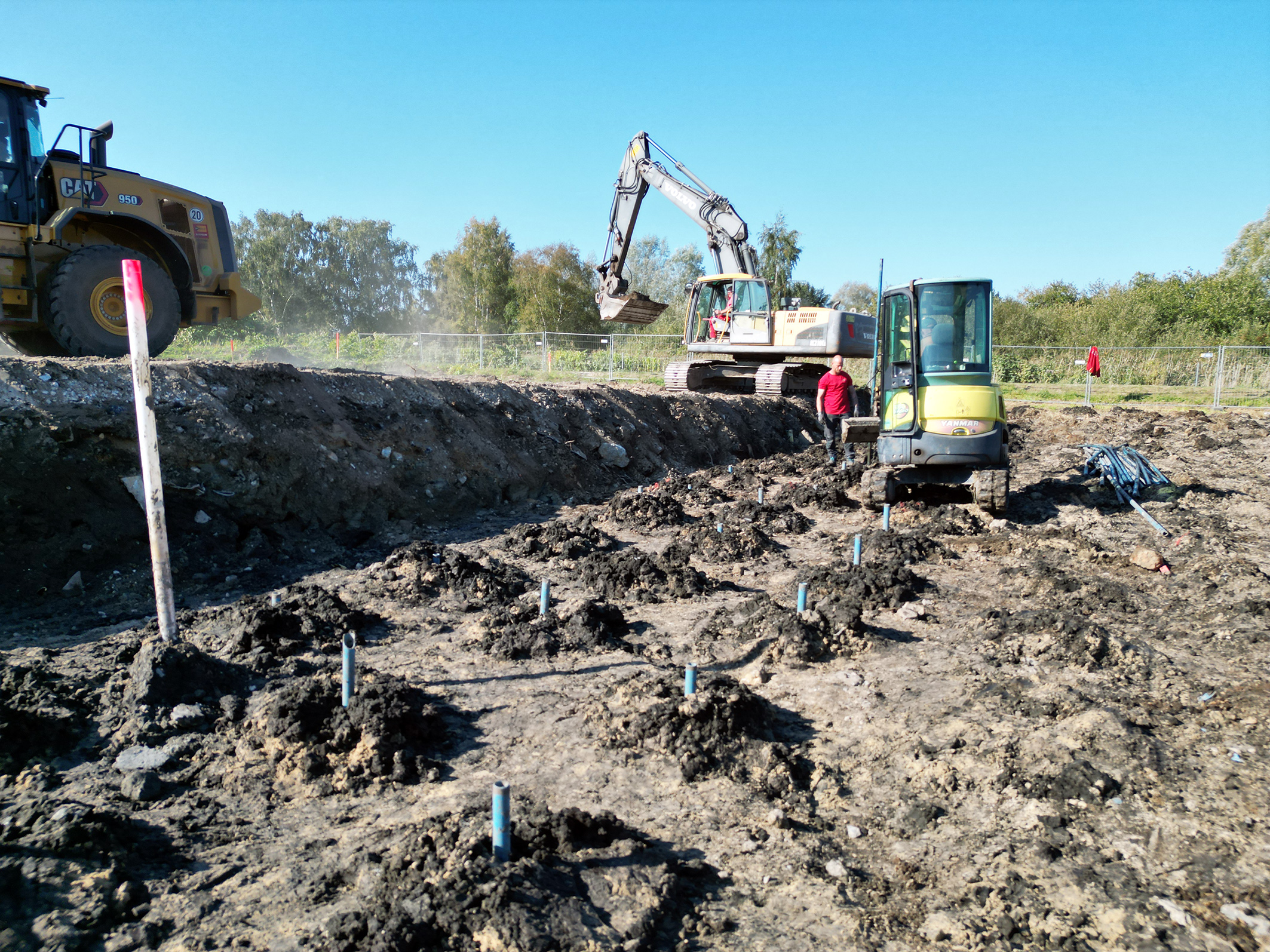 Baugrube mit mehreren blauen Markierungsstäben im Boden, Bagger und Radlader im Einsatz, Arbeiter in rotem Hemd auf der Baustelle