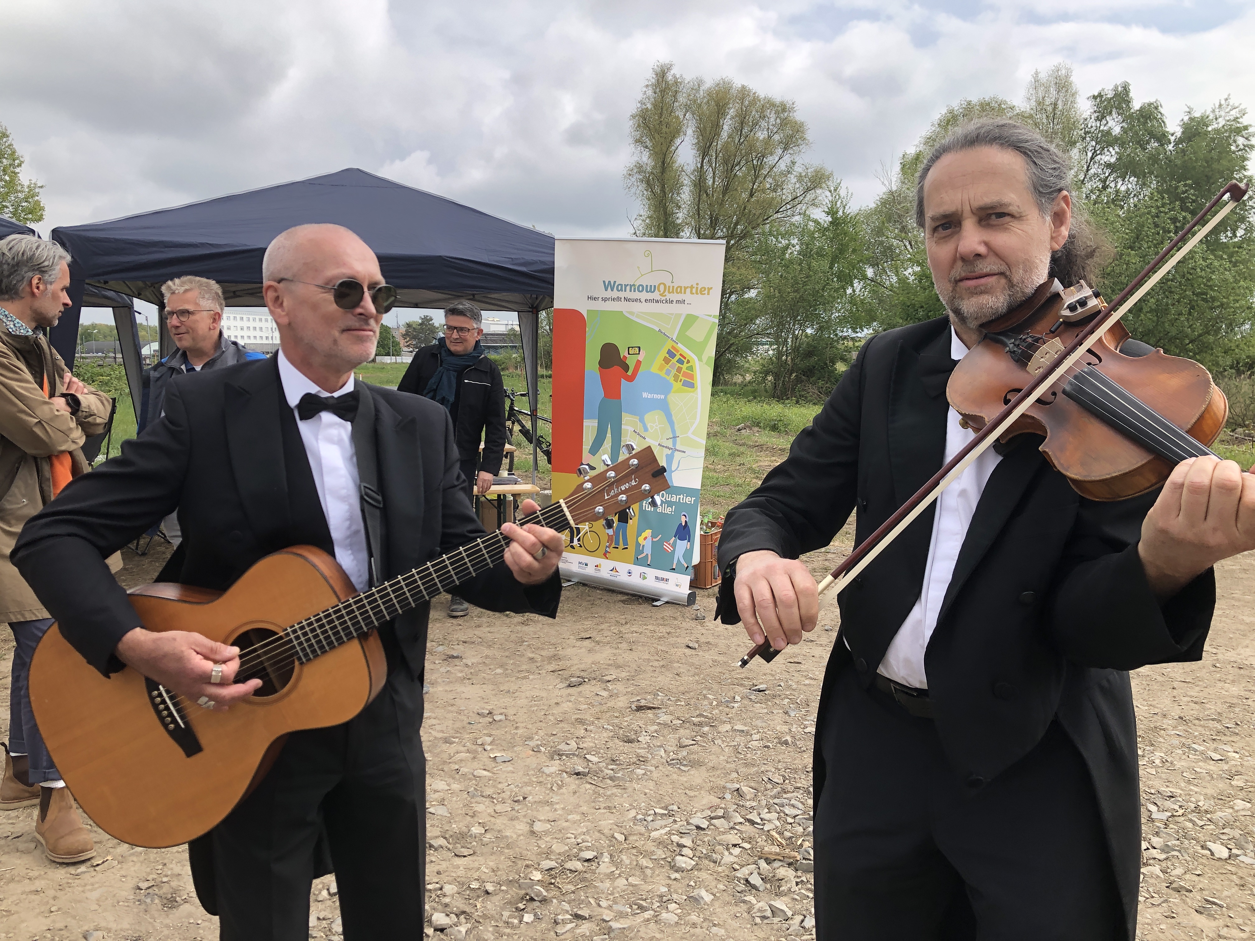 Zwei Musiker spielen im Freien, einer mit Gitarre, der andere mit Geige, umgeben von Bäumen